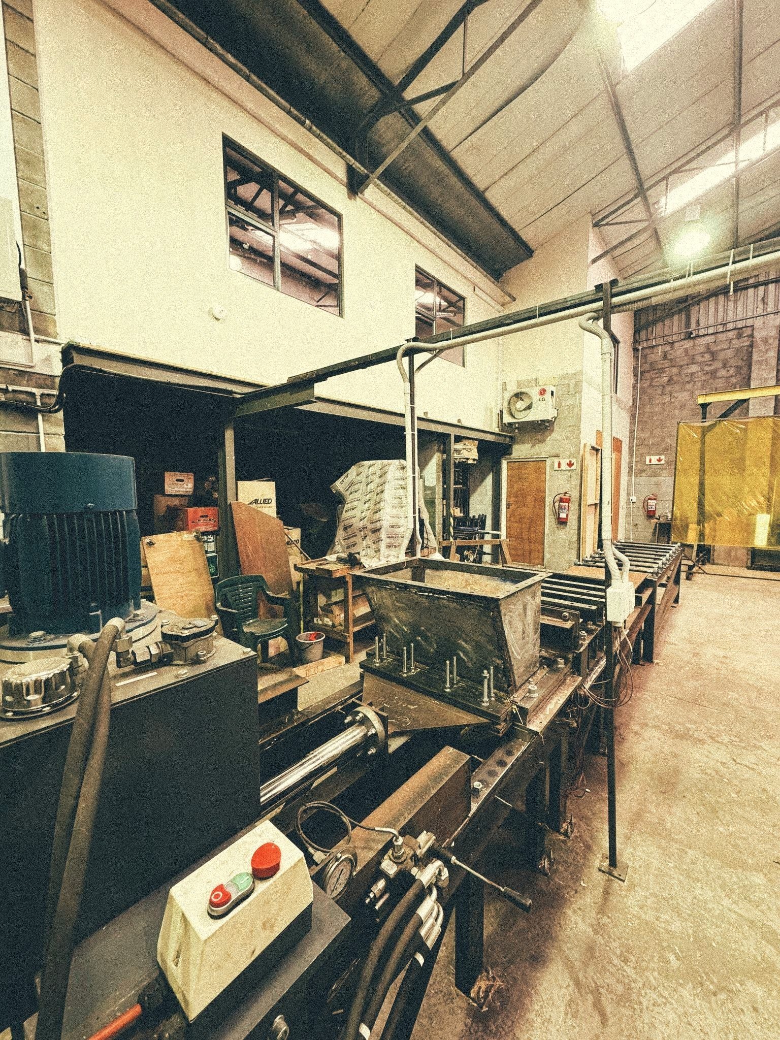 Prototype production line on the Bara workshop floor — long steel-framed conveyor with a feed hopper, electric drive motor on the left and a control station with red emergency-stop buttons in the foreground.