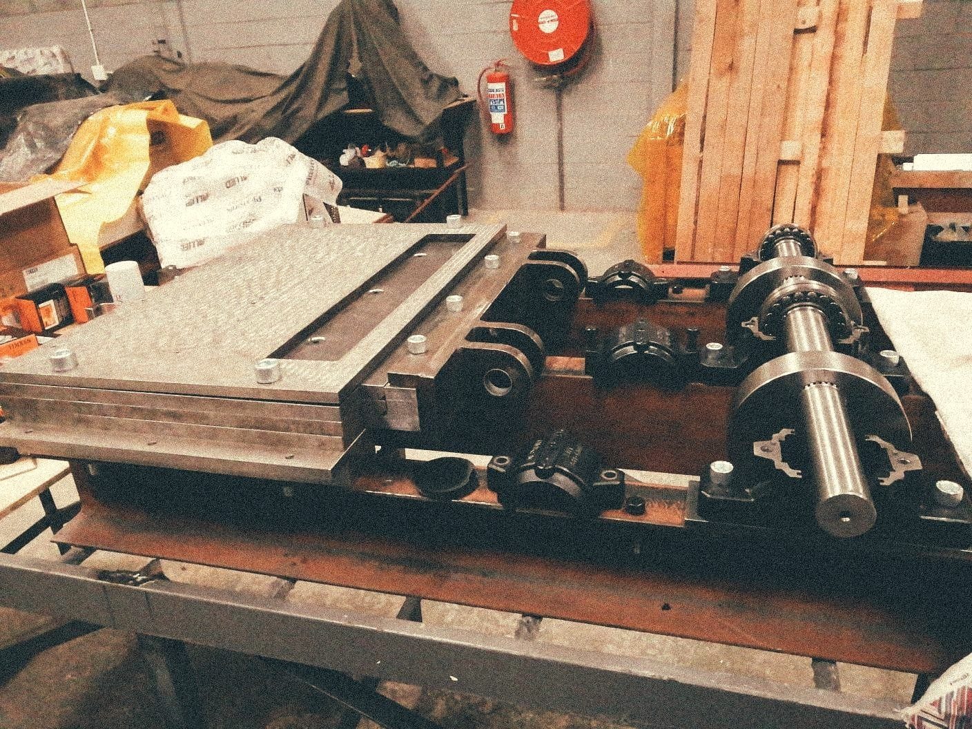 Heavy compression-roller assembly and drive shaft laid out on a steel fabrication bench in the Bara workshop — chain sprockets, bearings and tooled steel ready for assembly.