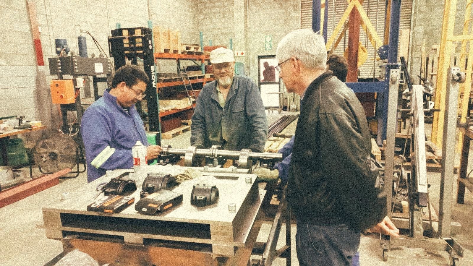 Three Bara engineers around a steel workbench fitting the drive-shaft and roller-bearing assembly together, the workshop hydraulic press and steel stock visible behind them.